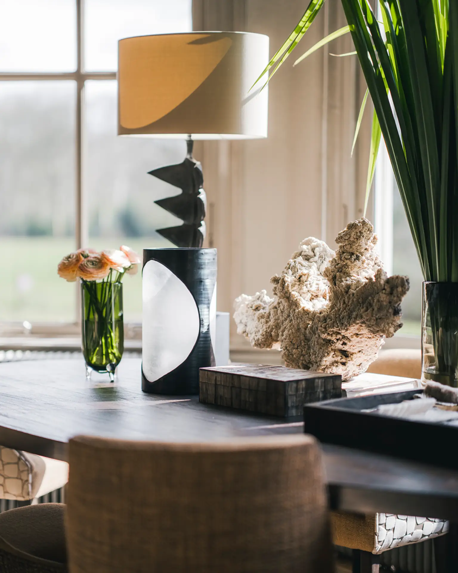 Luxe interieurdesign met gouden lamp, zwarte sculptuur en bloemen op donkere tafel bij raam